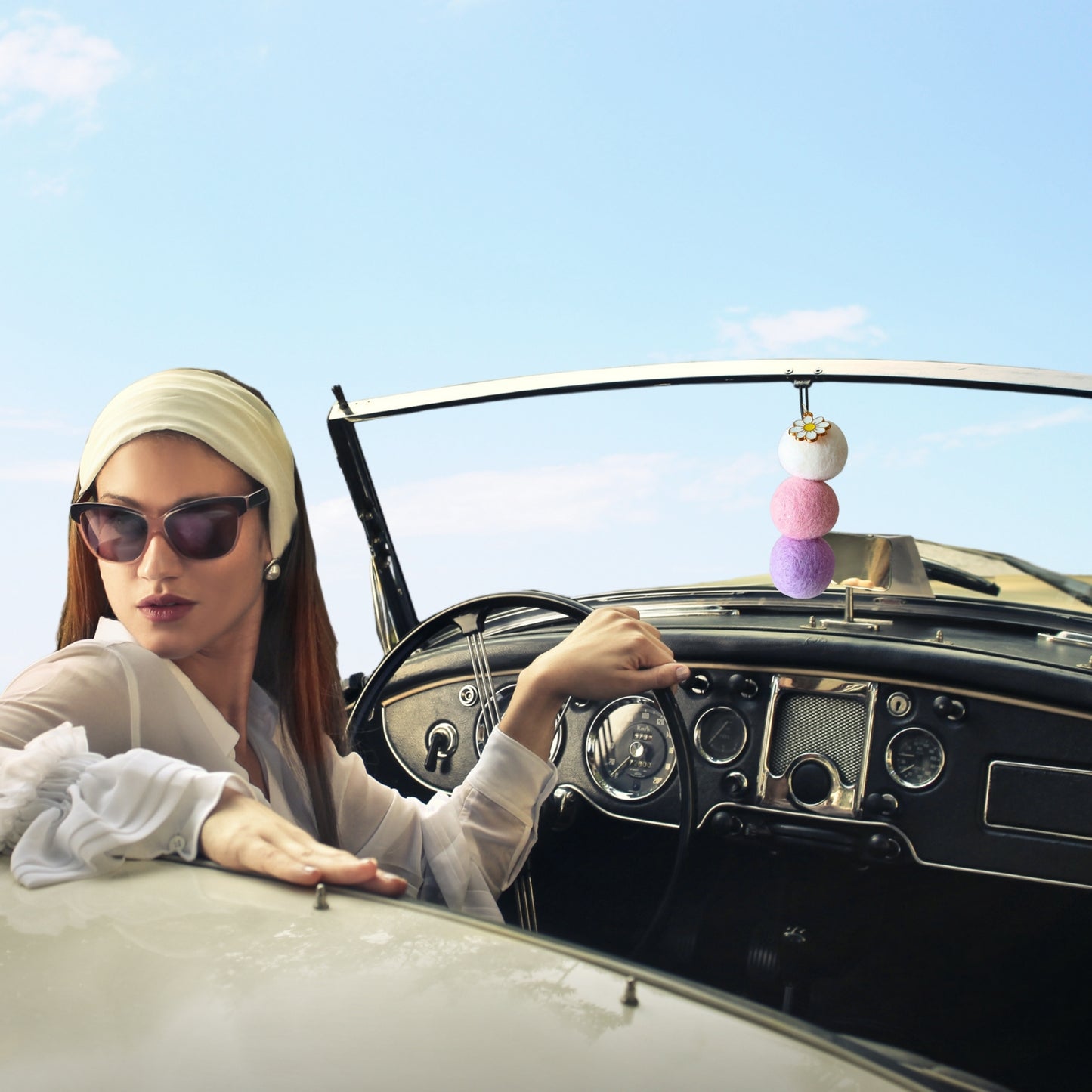 Woman sitting in a vintage car with a colorful car accessory hanging from the rearview mirror.