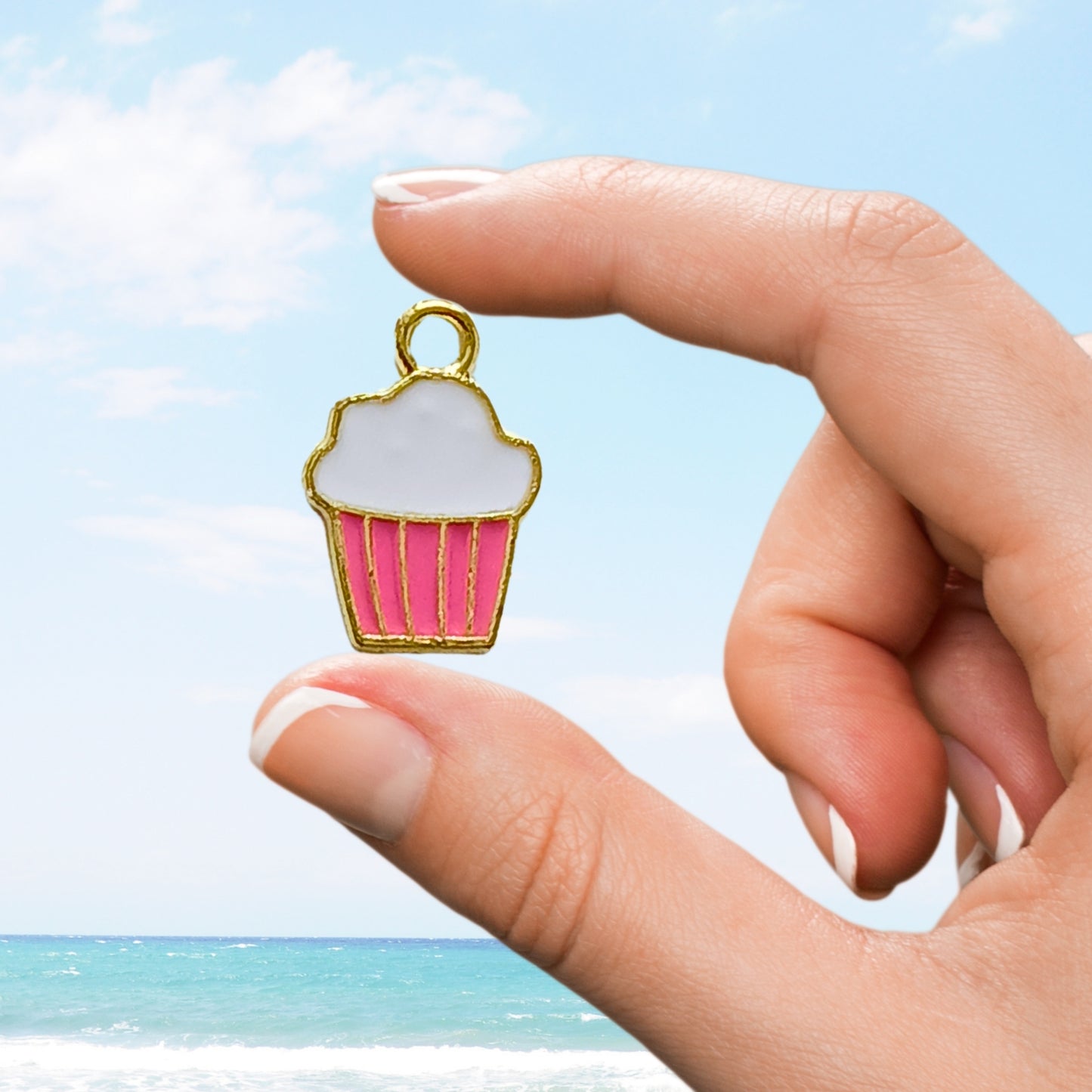 Hand holds pink cupcake charm with white icing above beach backdrop