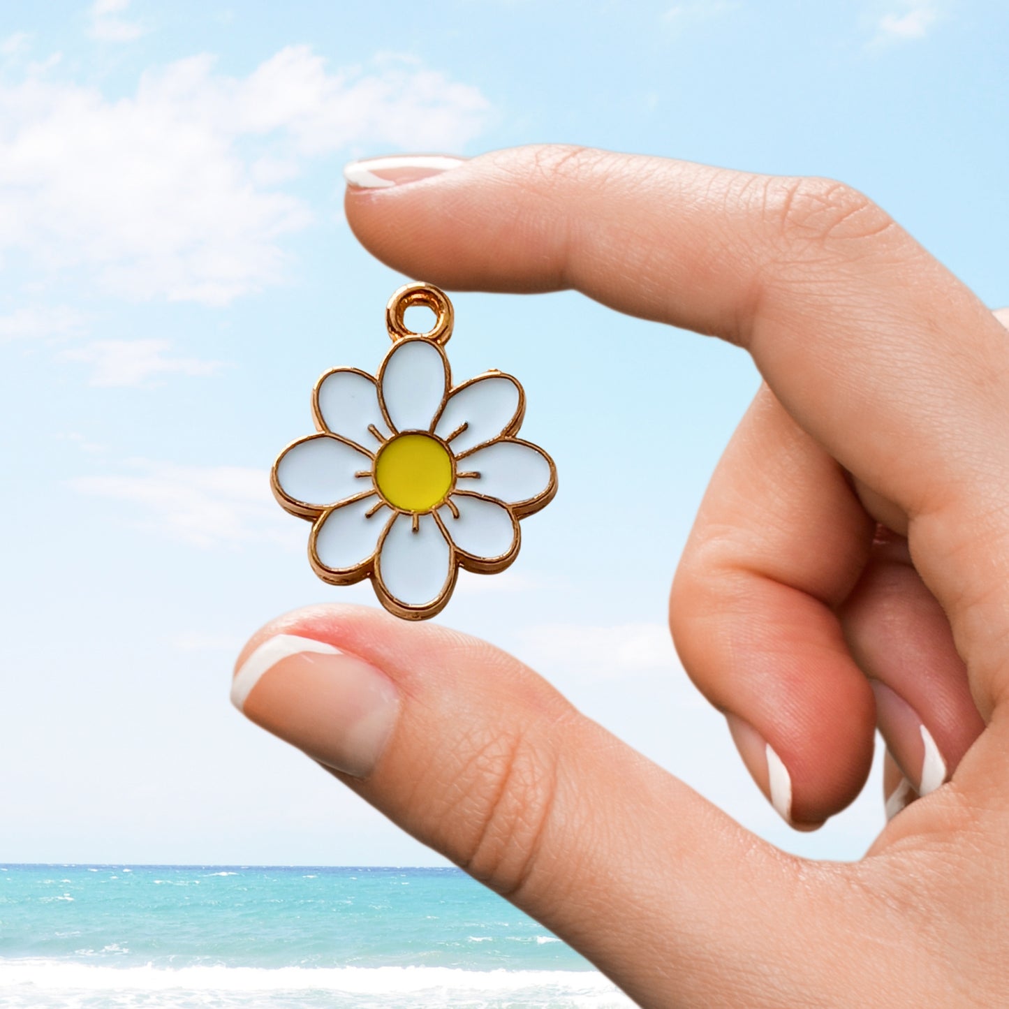 White daisy enamel charm held in fingers with yellow centre and ocean background
