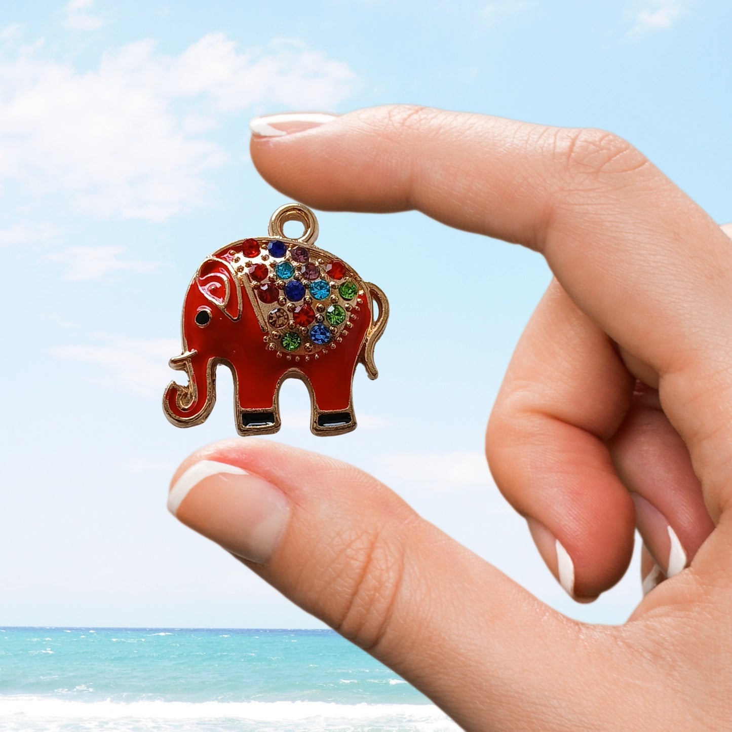 Hand holds red elephant charm with colourful gems on back by seaside
