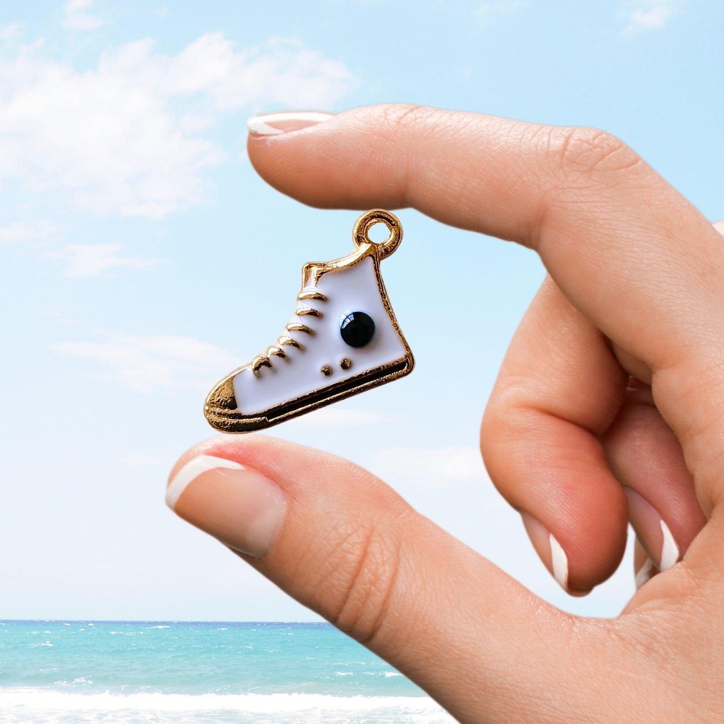 Hand holding a white high-top sneaker charm with black dot details over a beach scene