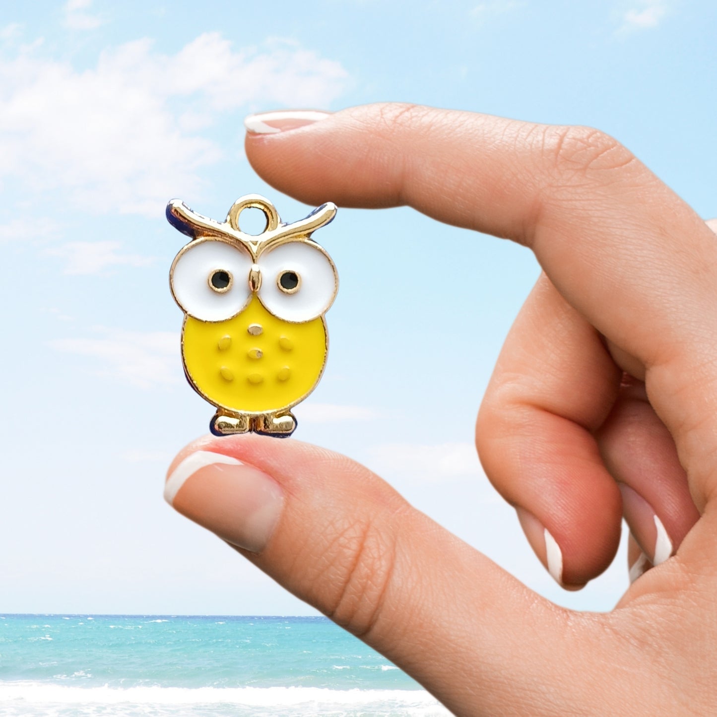 Hand displaying a yellow owl enamel charm by the seaside