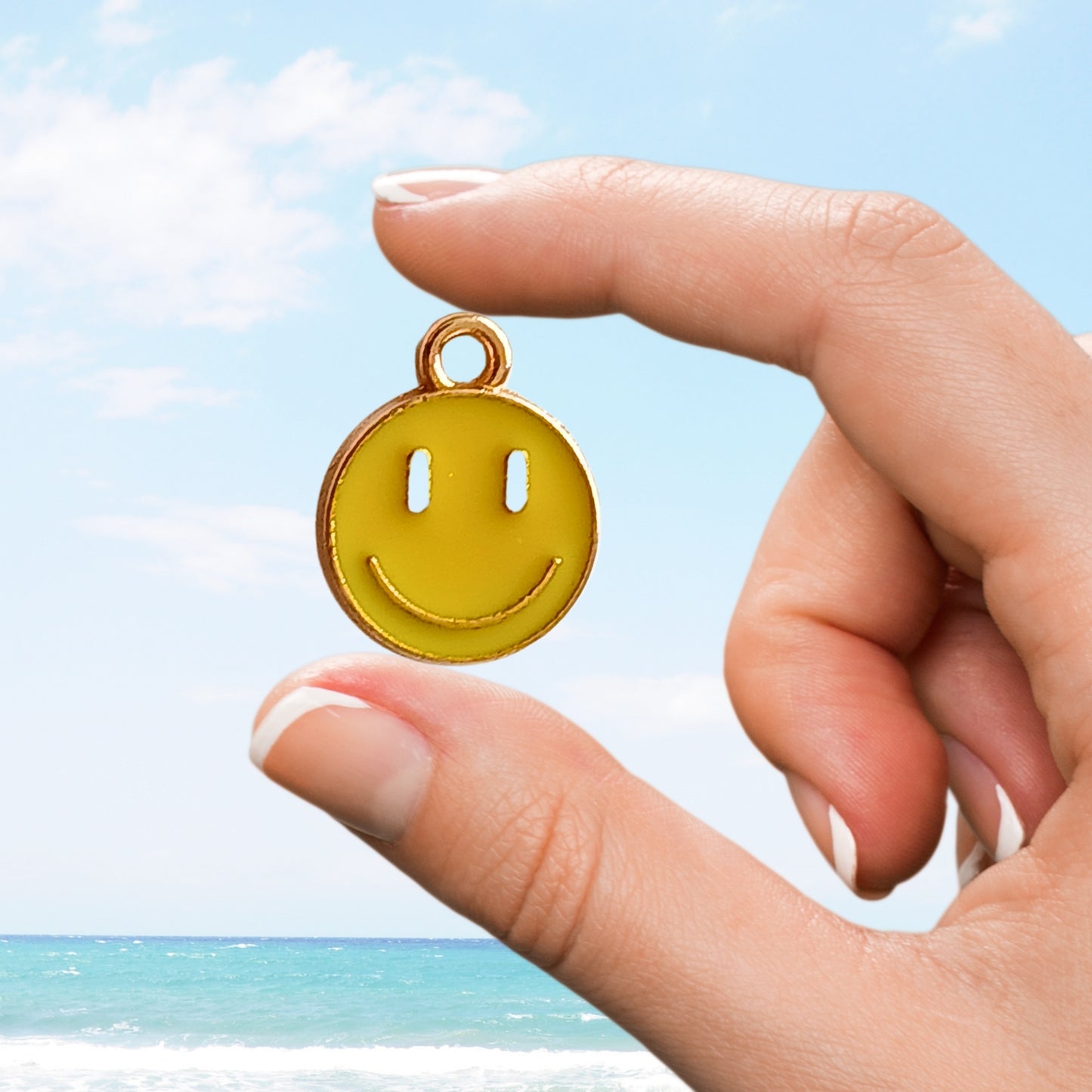 Yellow smiley face enamel charm pinched between fingers at the beach.