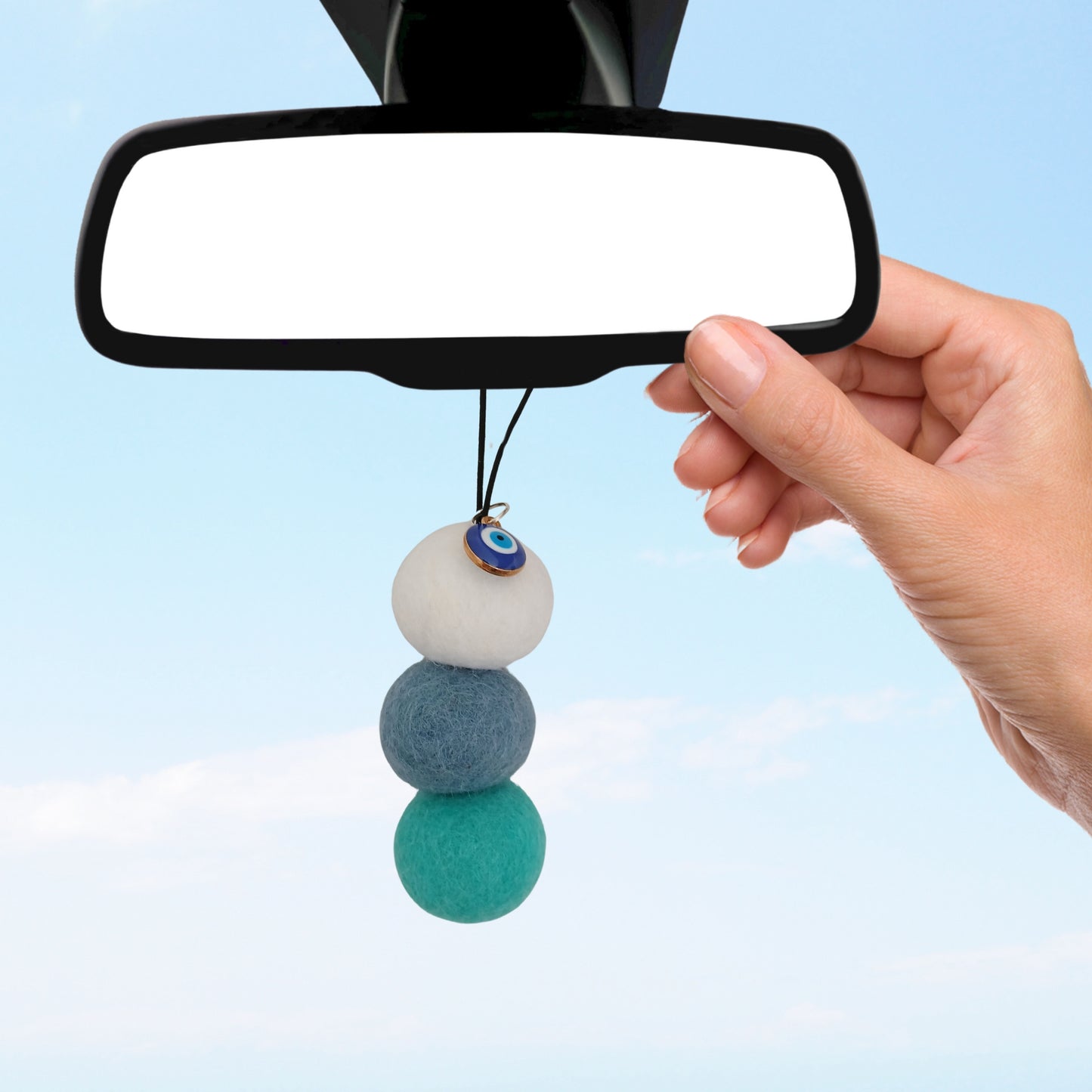 Hand hangs blue-toned felt ball car diffuser with evil eye charm from rearview mirror against bright sky.
