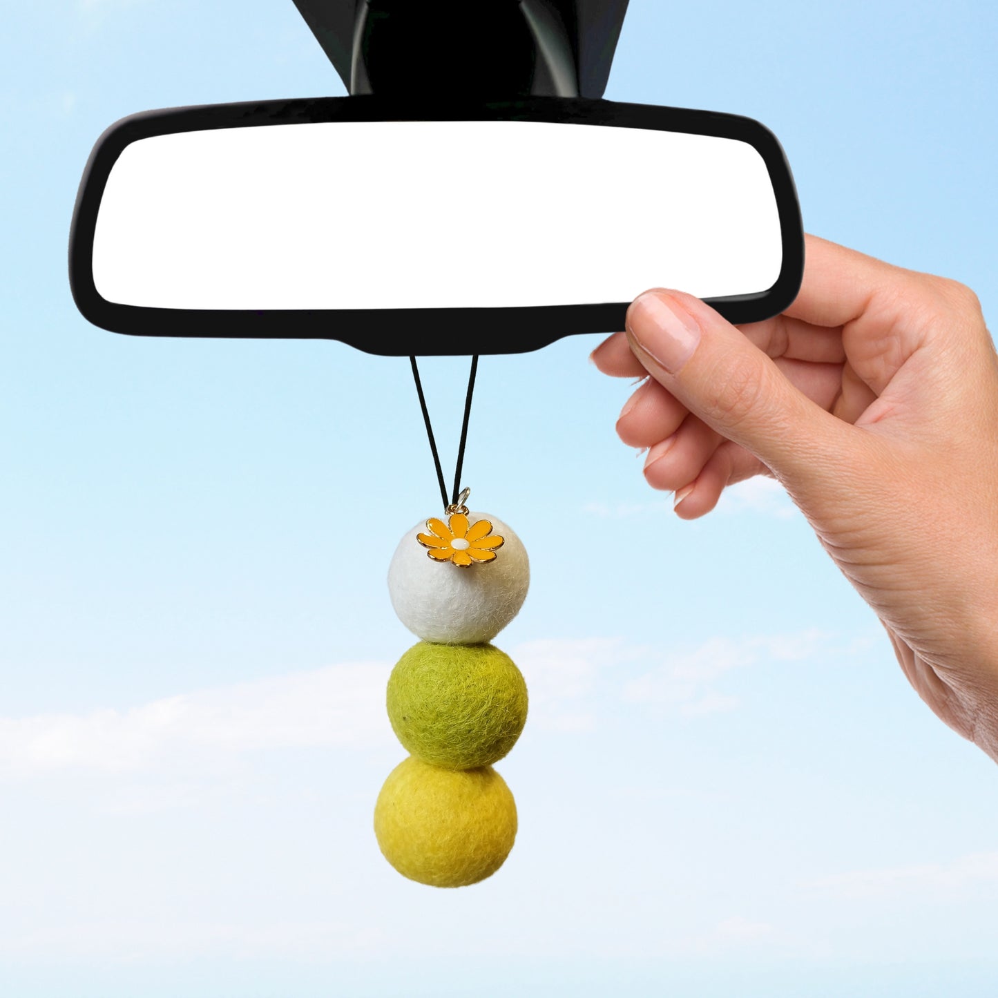 Person hanging a yellow and white felt ball diffuser with flower charm from a car’s rearview mirror, showcasing an eco-friendly car scent solution