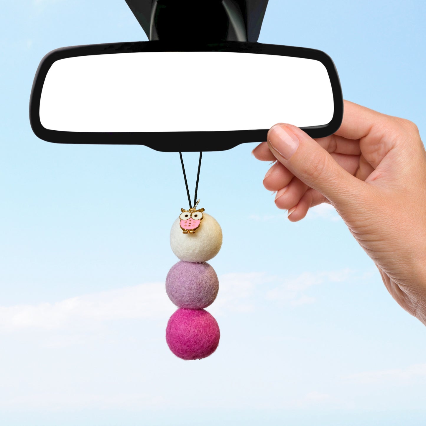 Hand hanging pink owl car diffuser with purple and white felt balls on rearview mirror against blue sky.