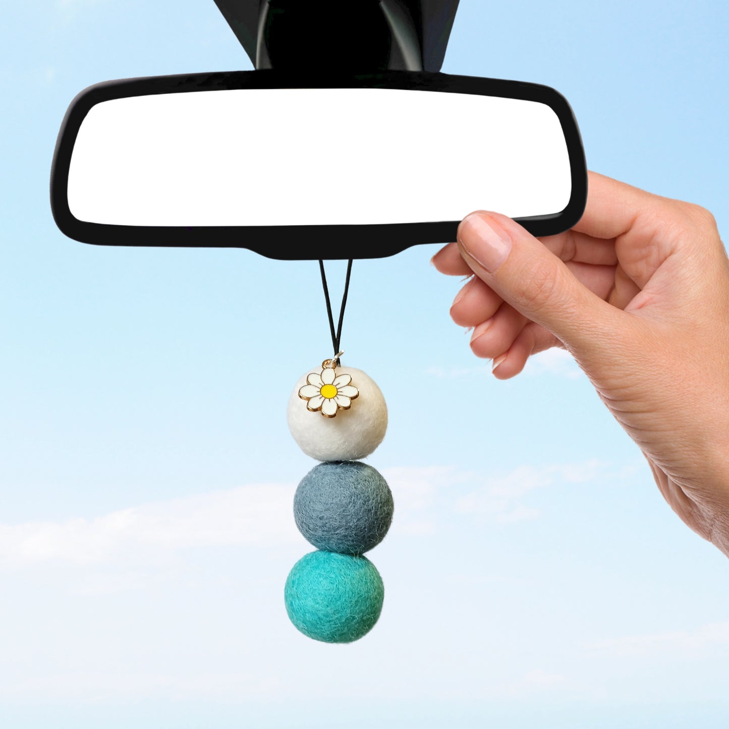 Hand installing blue and white felt ball car diffuser with daisy charm on rearview mirror, promoting a reusable and stylish car air freshener alternative