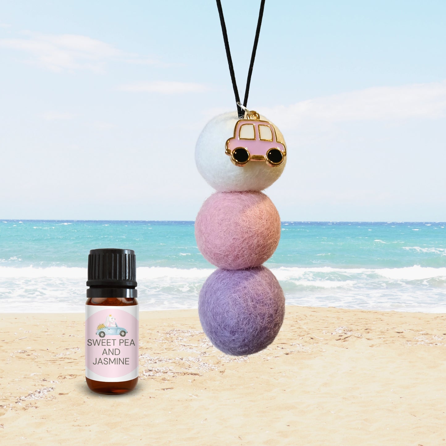 Sweet Pea & Jasmine car diffuser with pastel felt balls and pink enamel car charm, displayed on beach background with fragrance bottle