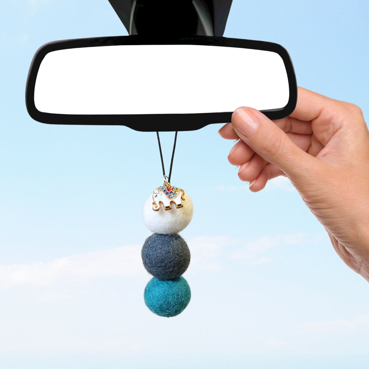Hand hanging a three-ball wool felt car diffuser with elephant charm on rear-view mirror, photographed against a blue sky.