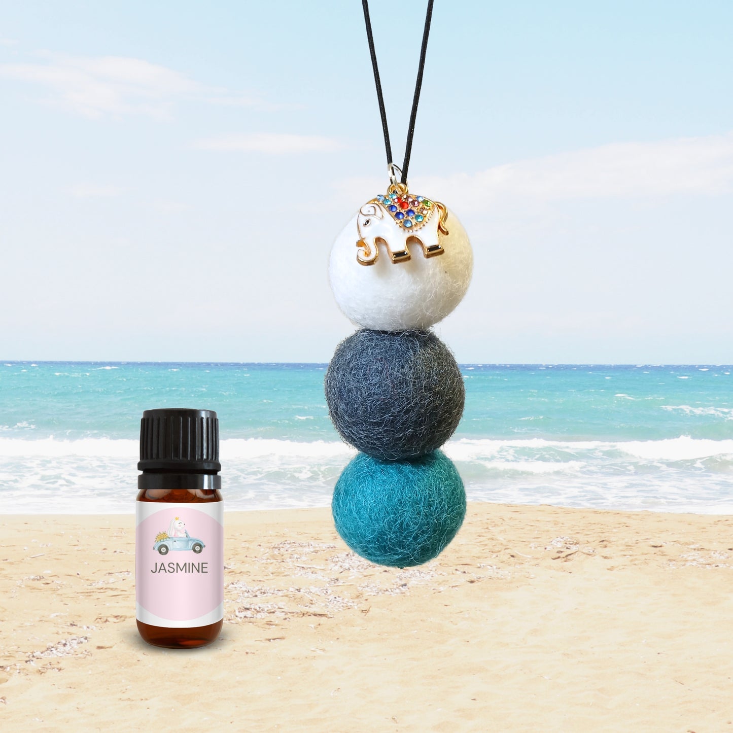 Eco-friendly felt ball car diffuser with white, navy, and teal wool balls, enamel elephant charm, and jasmine fragrance oil bottle on a beach background