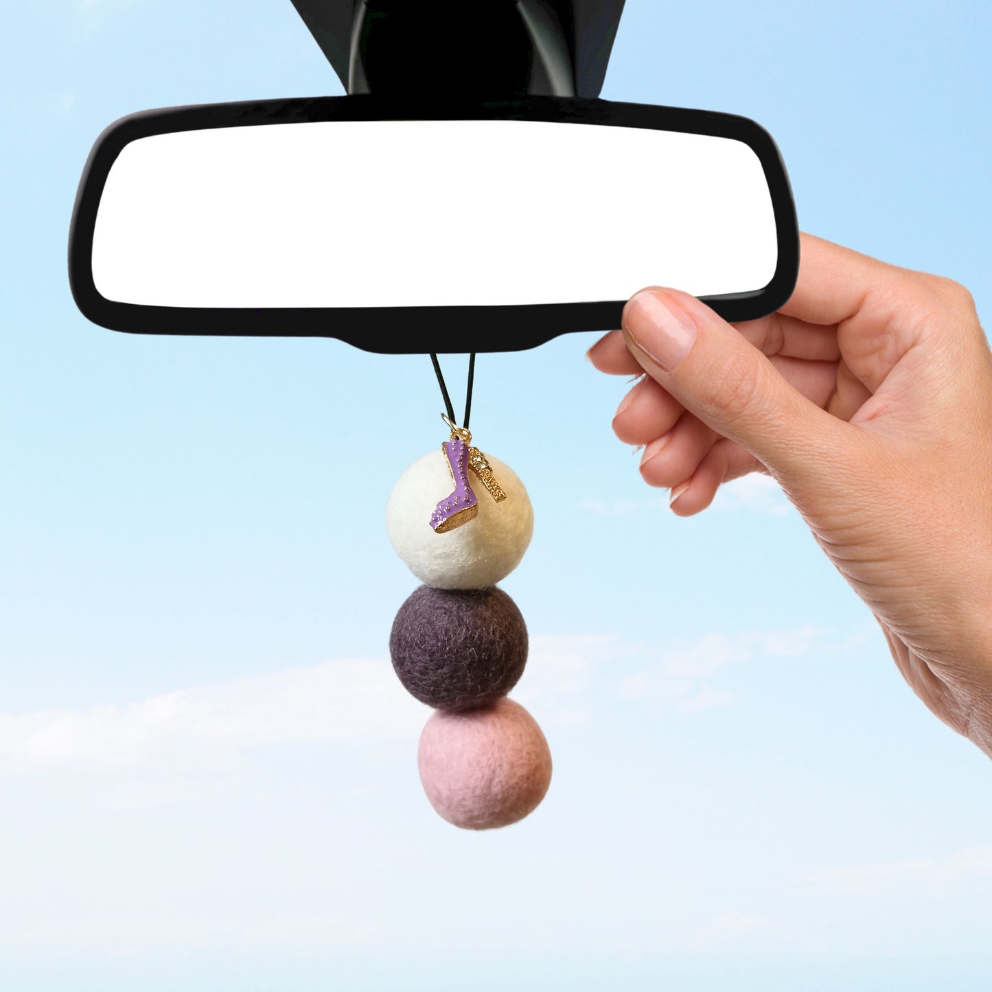 Hand hangs a purple boot charm felt ball diffuser from a car's rearview mirror with a blue sky background.