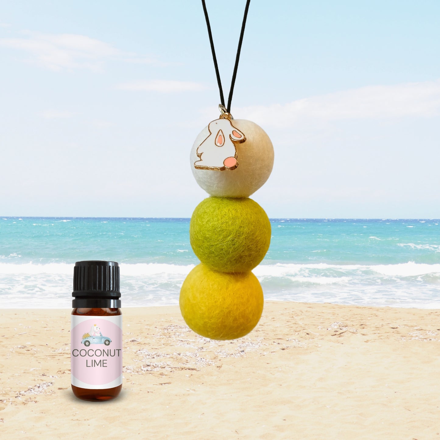 Coconut Lime car diffuser with bunny charm and yellow-green felt balls displayed on a beach next to a fragrance oil bottle