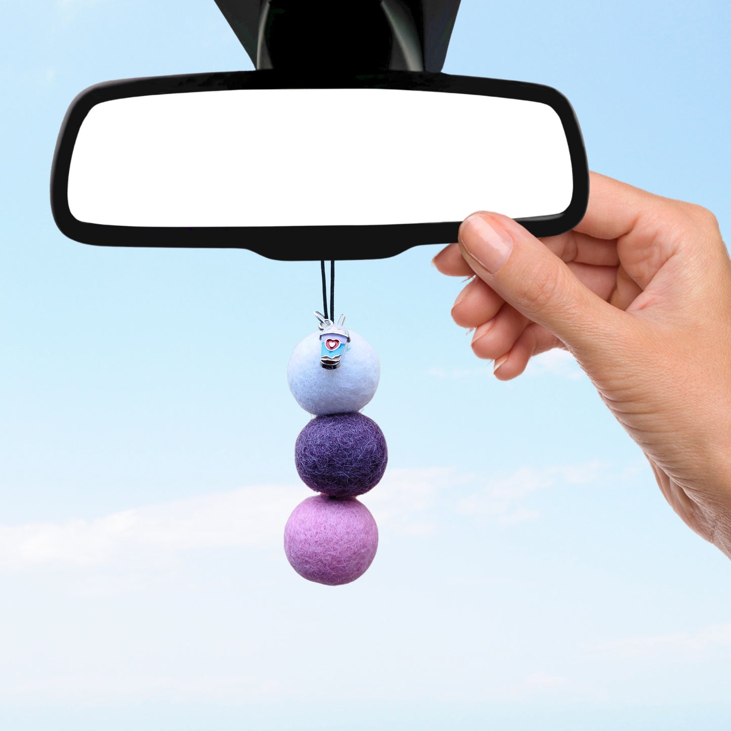 Hand hangs pastel felt ball car diffuser with charm from rearview mirror.