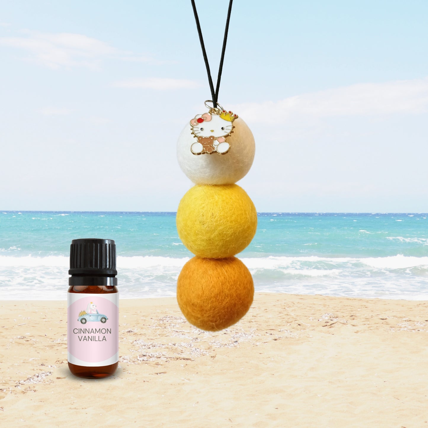 Eco-friendly car diffuser with white, yellow, and orange felt balls, cute enamel charm, and cinnamon vanilla fragrance oil on sandy beach backdrop