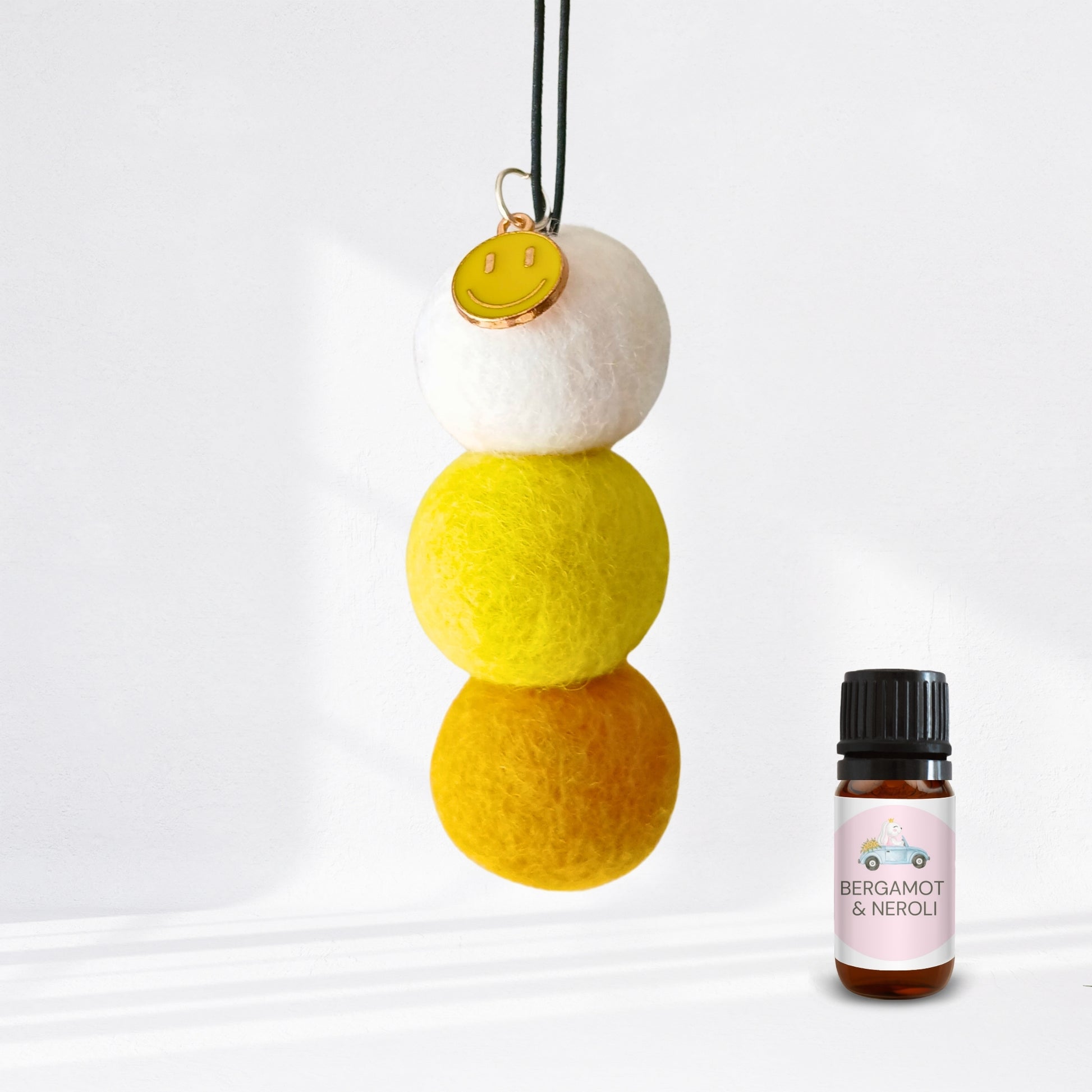 Three-tiered felt ornament with a yellow smiley face charm next to a bottle of Bergamot & Neroli essential oil.