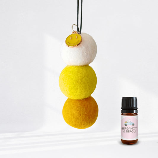 Three-tiered felt ornament with a yellow smiley face charm next to a bottle of Bergamot & Neroli essential oil.
