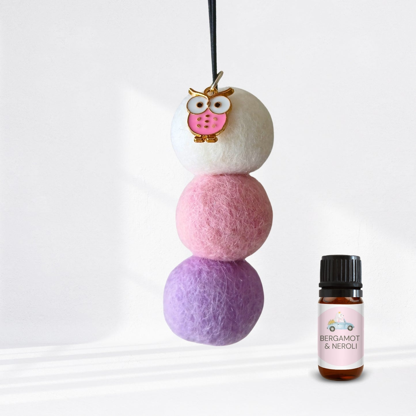 Stack of colorful felt balls with a small owl charm next to a bottle labeled 'Bergamot & Neroli' on a white background