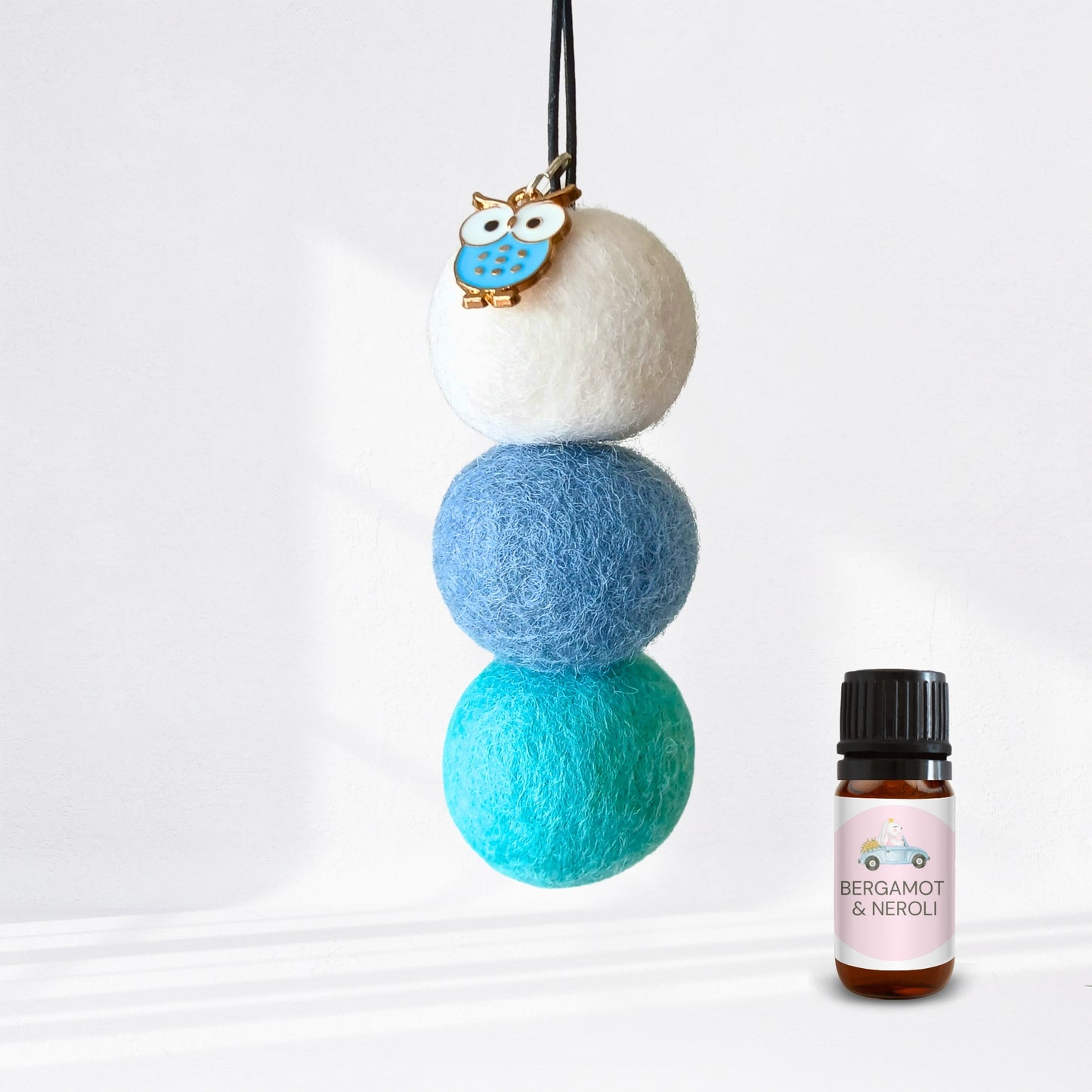 Three felted balls in white, blue, and teal with a small owl charm, next to a bottle of Bergamot & Neroli essential oil.