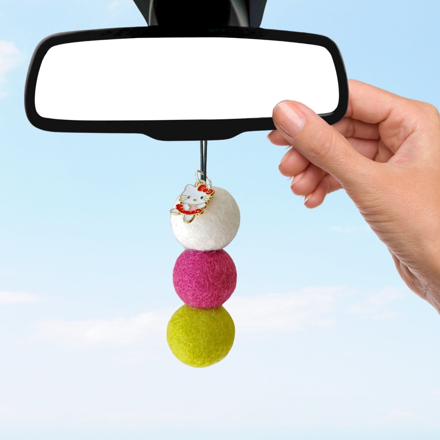 Colorful felt ball car charm attached to a car's rearview mirror against a clear blue sky.