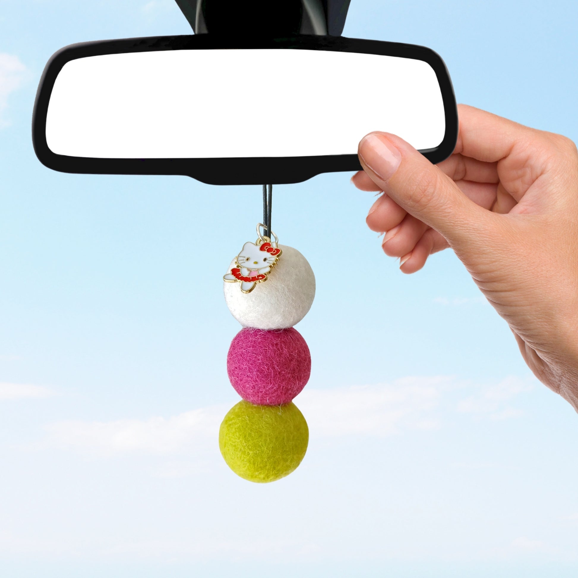 Colorful felt ball car charm attached to a car's rearview mirror against a clear blue sky.