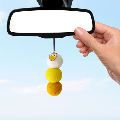 Hand hanging a cartoon character on felt balls hanging from a car mirror against a clear blue sky.