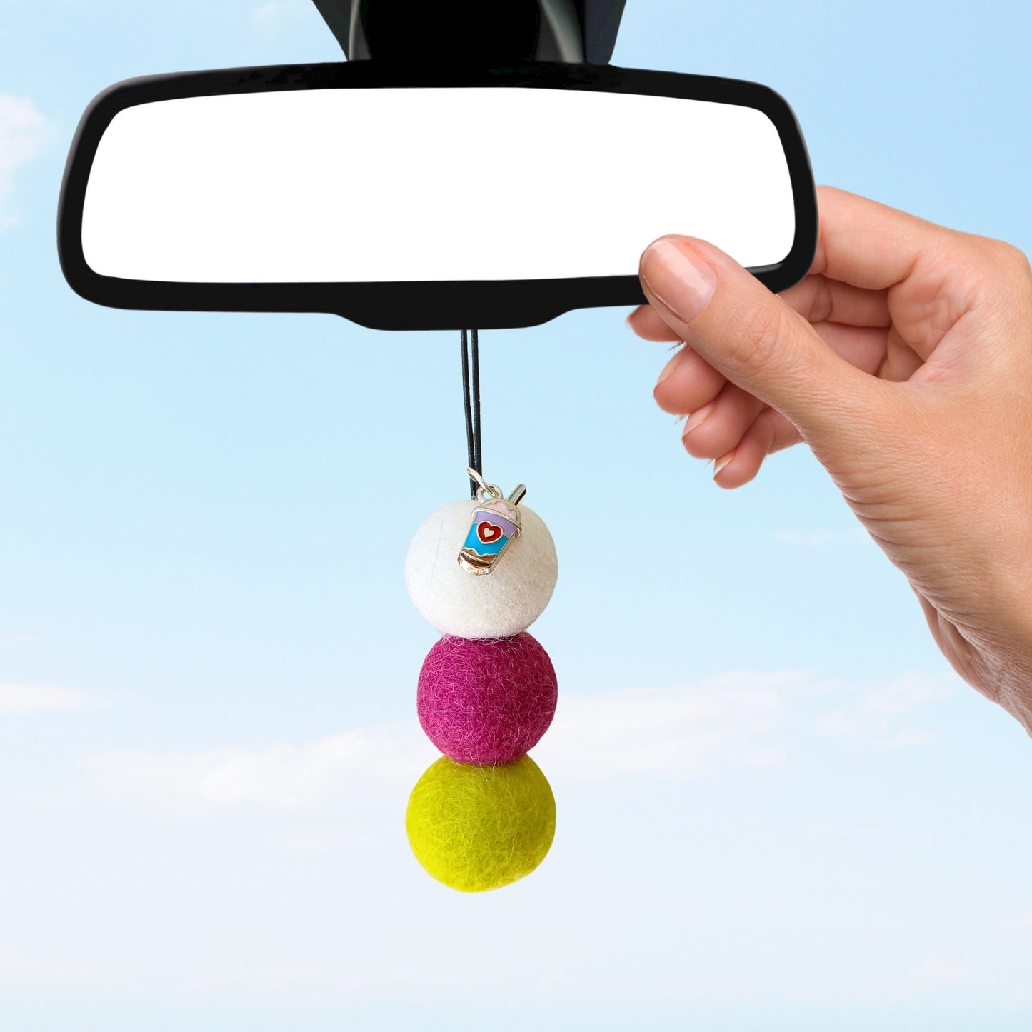 Colorful pom-pom car charm being attached to a car's rearview mirror against a clear blue sky.