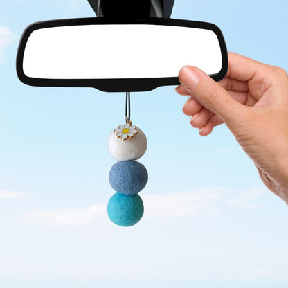 Hand holding a blue and white decorative car charm in front of a car's rearview mirror.