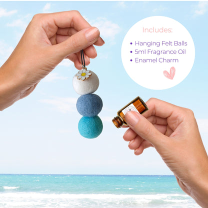 hanging car diffuser with felt balls and a small bottle of fragrance oil against a beach background