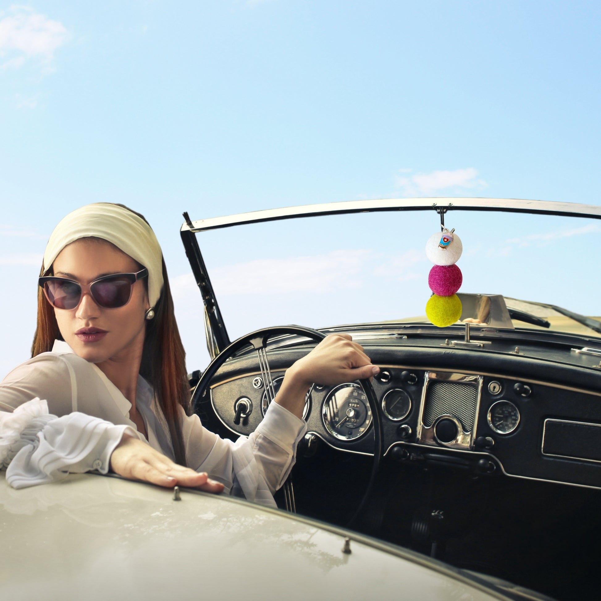 Woman sitting in a vintage car with a colorful car accessory hanging from the rearview mirror.