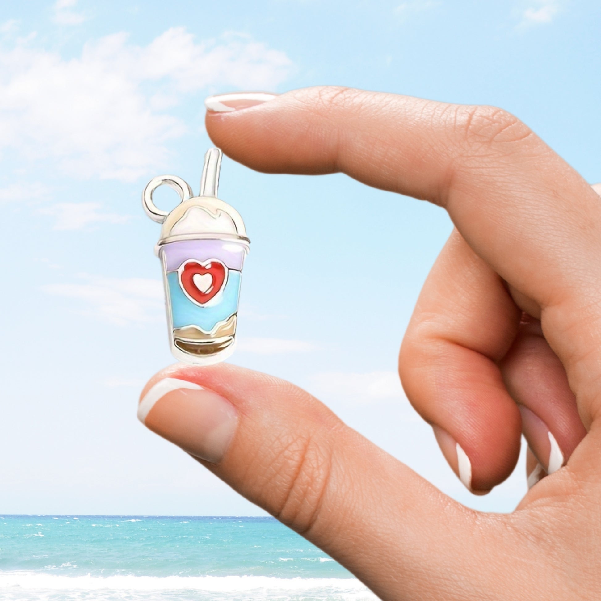 Hand holding a small cup-shaped charm with a heart design against a beach background