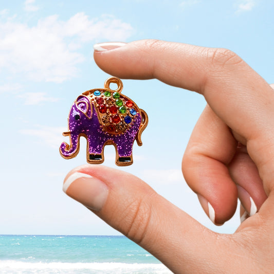 Hand holding a glittery purple elephant charm with rainbow gems and gold outline in front of ocean backdrop