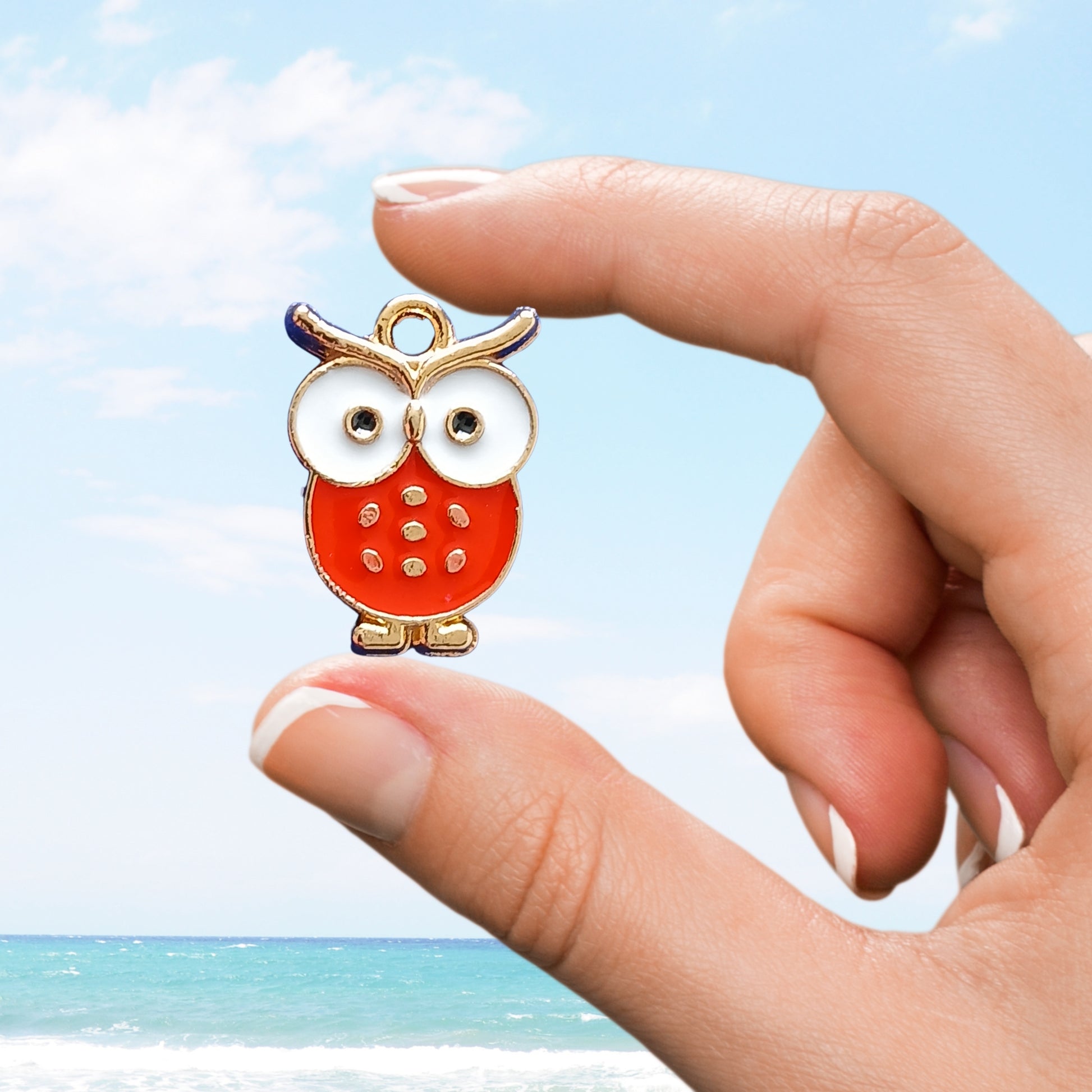 Hand holding a red owl enamel charm against a beach background.