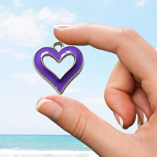 Hand holding a silver heart charm with a bold purple enamel outline against a seaside background
