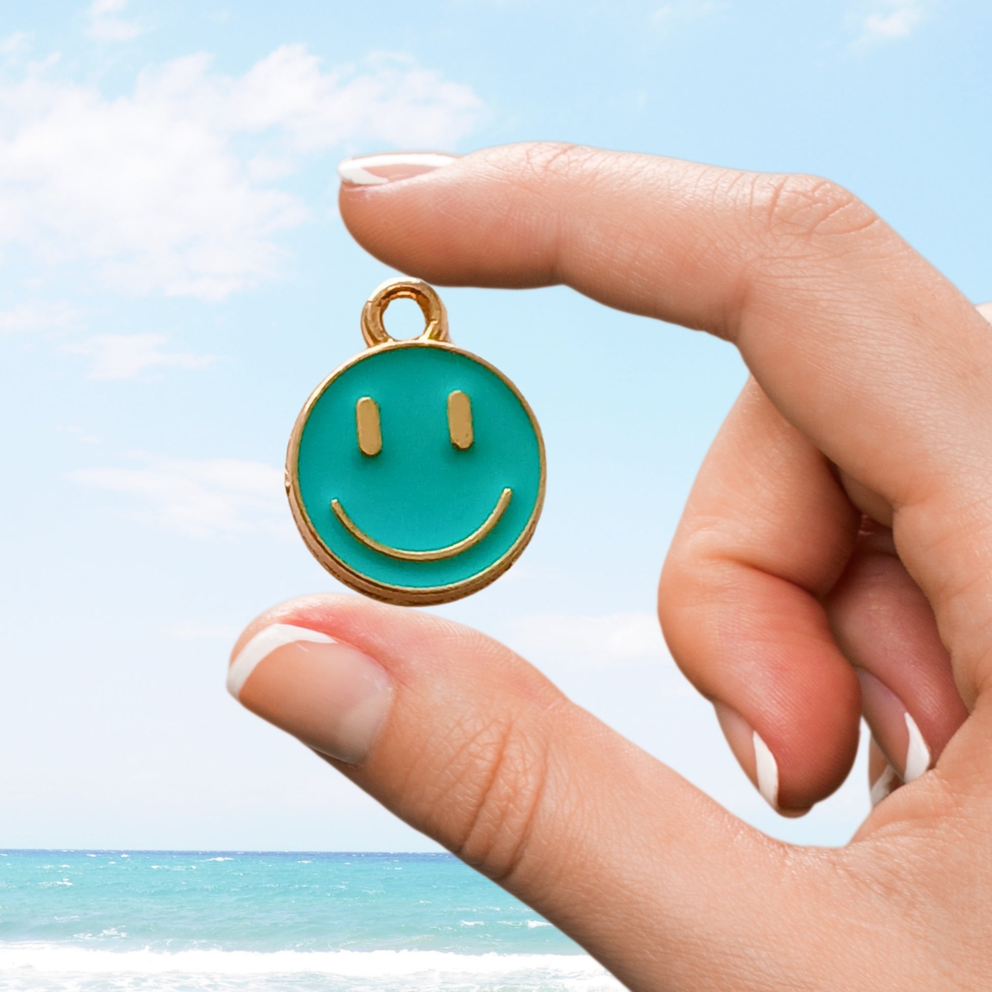 Teal smiley face charm held between fingers with ocean backdrop
