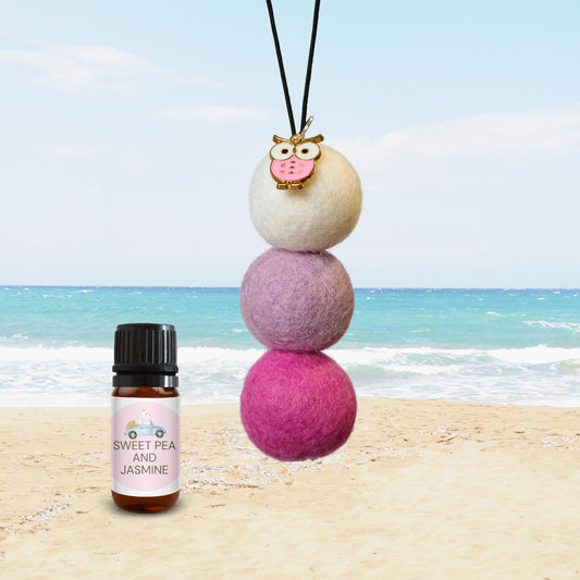 Car diffuser with pink owl charm and pastel felt balls beside Sweet Pea & Jasmine essential oil bottle on beach background