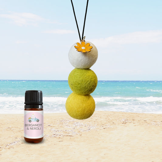 Natural wool felt ball car diffuser with yellow flower charm and Bergamot & Neroli essential oil bottle shown on a beach, highlighting refillable, non-toxic air freshening