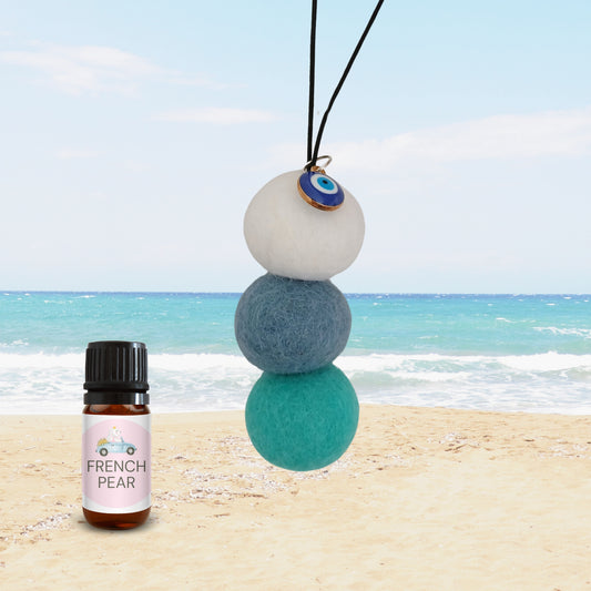 Hanging car diffuser with white, blue, and teal felt balls, evil eye enamel charm, and French Pear fragrance oil bottle on sandy beach background.