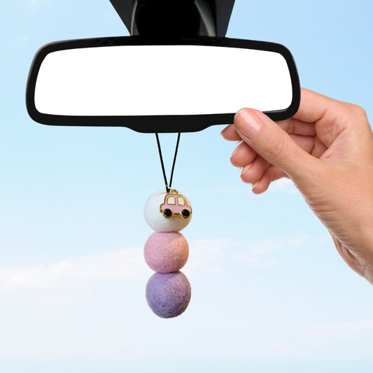 Hand hanging Sweet Pea & Jasmine pastel car diffuser with pink car charm from rearview mirror against blue sky.