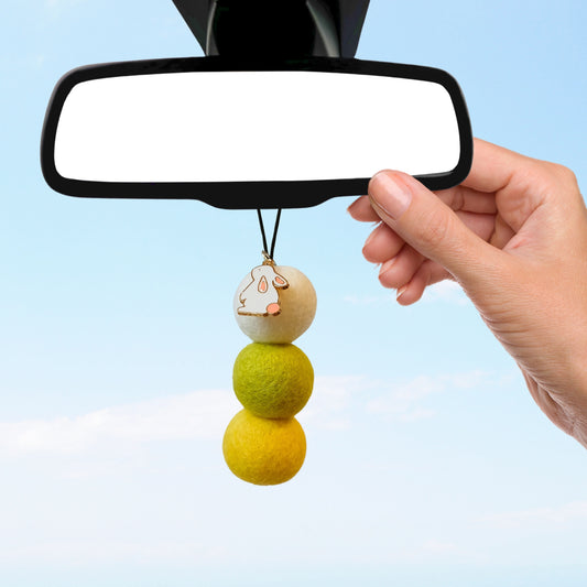 Hand hangs Coconut Lime bunny car diffuser on rearview mirror with blue sky background.