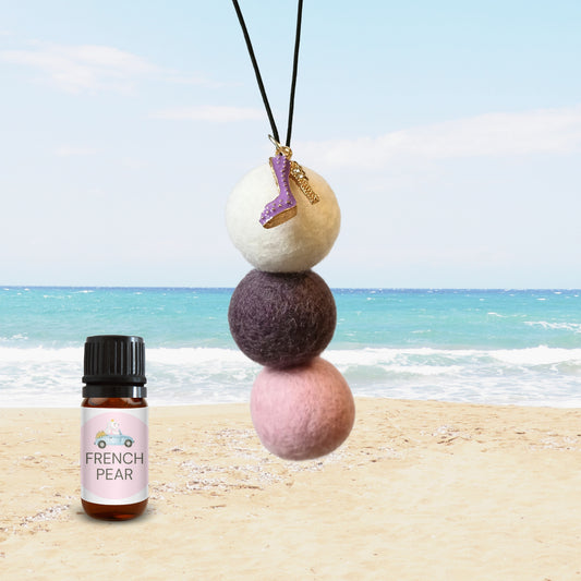 Close-up of a car air freshener with three stacked felt balls in white, plum, and blush pink, featuring a purple high-heel enamel charm, paired with a French Pear fragrance oil bottle on a beach backdrop.